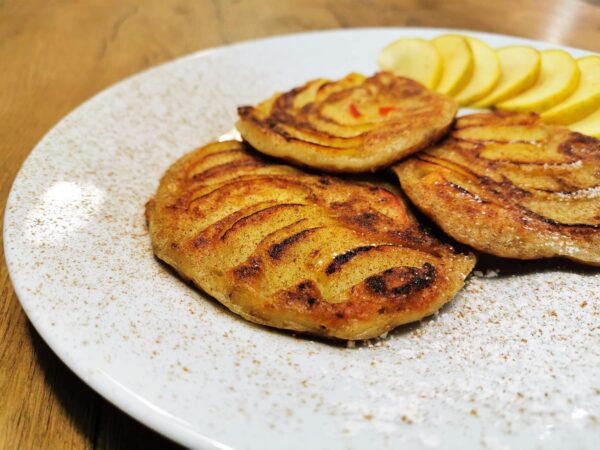 Drei Apfelpancakes serviert auf einem weißen Teller, garniert mit frischen Apfelscheiben und bestreut mit Zimt.