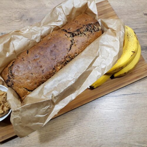Bananen-Nuss-Schokokuchen rundherum eingepackt in Backpapier, angerichtet auf einem Holzbrett mit einer weißen Schale gefüllt mit Erdnussbutter und einem Teelöffel, garniert mit einem Bund frischer Bananen.