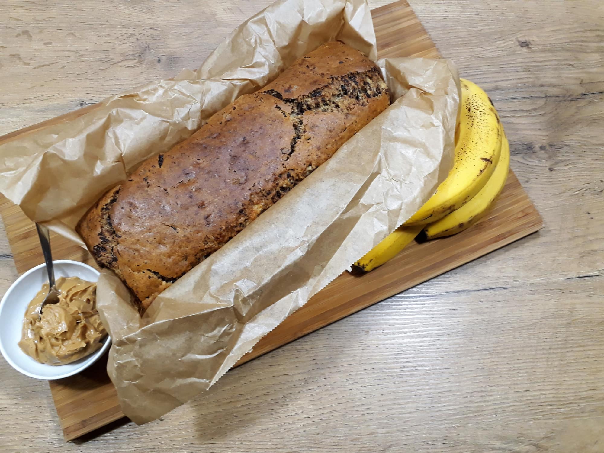 Bananen-Nuss-Schokokuchen rundherum eingepackt in Backpapier, angerichtet auf einem Holzbrett mit einer weißen Schale gefüllt mit Erdnussbutter und einem Teelöffel, garniert mit einem Bund frischer Bananen.