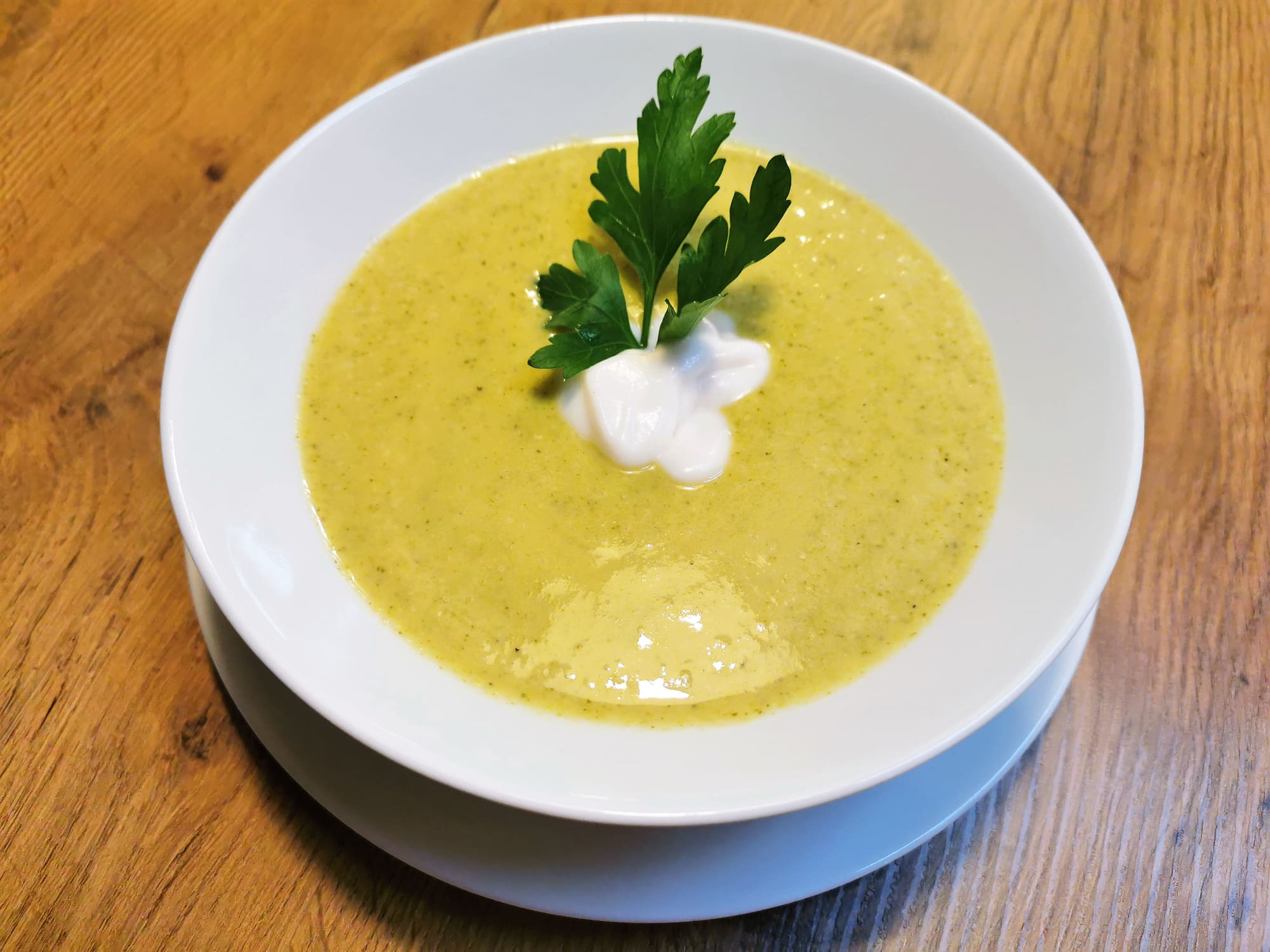 Brokkolicremesuppe serviert in einem weißen Suppenteller, garniert mit einem Klecks Pflanzenjoghurt und einem kleinen Petersilienzweig.