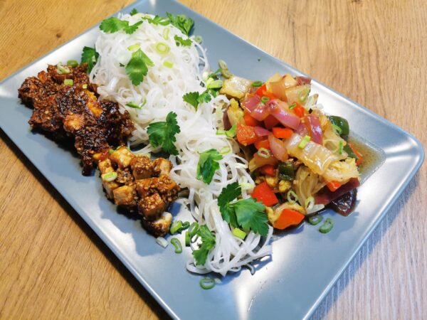 Chinakohl-Asia-Pfanne mit Reisnudeln und Sesam-Tofu angerichtet auf einem blauen Teller, garniert mit Petersilienblättern.
