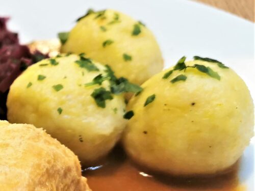 Erdäpfelknödel als Beilage zu einem Braten, mit Bratensauce und Rotkraut, serviert auf einem weißen Teller.