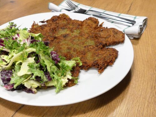 Ein Erdäpfelwatsch serviert auf einem weißen Teller mit einem gemischten Blattsalat mit Pflanzenjoghurtdressing. Daneben liegt ein Essbesteck auf einer karierten Stoffserviette.