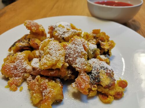 Kaiserschmarrn serviert auf einem weißen Teller, bestreut mit Staubzucker und einer kleinen Schale Zwetschkenröster im Hintergrund.