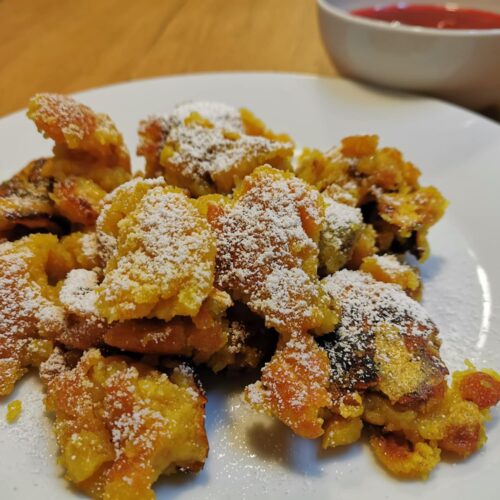 Kaiserschmarrn serviert auf einem weißen Teller, bestreut mit Staubzucker und einer kleinen Schale Zwetschkenröster im Hintergrund.