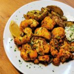 Karfiol-Wings und Kräutererdäpfeln mit Knoblauchsauce angerichtet auf einem weißen Teller, garniert mit einer Zitronenspalte und frisch gehackter Petersilie.