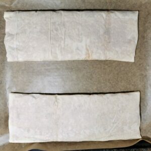 Zwei fertig gefüllte und gewickelte Krautstrudel auf einem mit Backpapier belegten Backblech, fertig um in den Ofen geschoben zu werden.