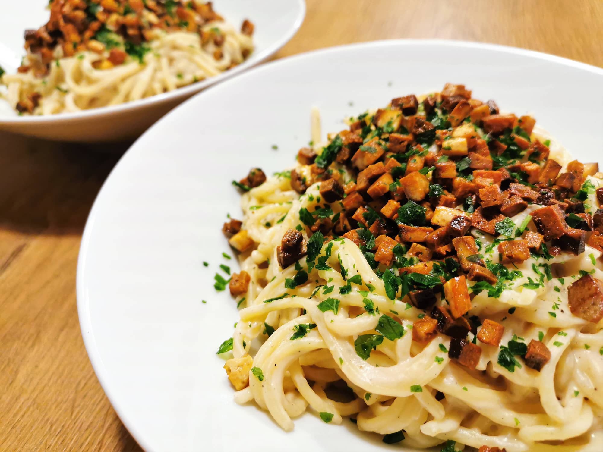 Spaghetti-Pasta mit Karfiolcreme, bestreut mit Tofuspeckwürfeln und gehackter Petersilie, angerichtet in einem weißen Teller.
