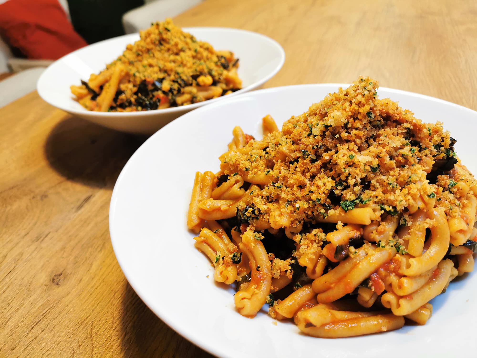 Pasta mit Schwarzkohl in Paradeisersauce, bestreut mit knusprigen Kräuterbröseln, angerichtet in weißen Tellern.