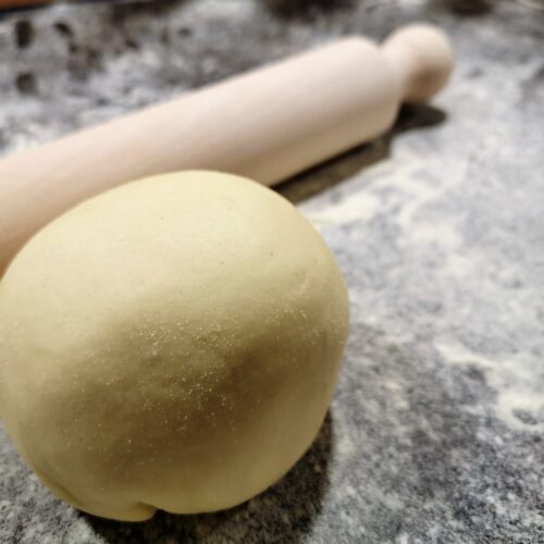 Eine Teigkugel frischer Pastateig auf einer bemehlten Arbeitsfläche mit einem Nudelholz.