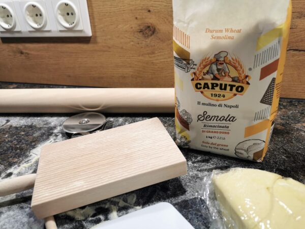 Eine bemehlte Arbeitsfläche mit einer geöffneten Packung Semola Pastamehl, einem Nudelholz, einer Teigkarte, einem Teigrad, einem geriffelten Nudelbrett und einem frischen Pastateig in Frischhaltefolie eingewickelt.