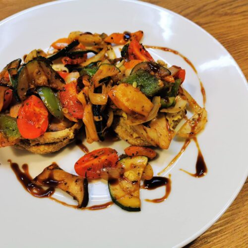 Pfannenbrot belegt mit Hummus und gebratenem mediterranem Gemüse, serviert auf einem weißen Teller und getoppt mit Crema di Balsamico.