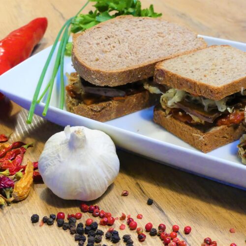 Drei Sandwiches auf einem weißen Servierteller, jedes Sandwich wurde mit einer anderen Brotsorte zubereitet. Der Teller ist mit frischen Kräutern, frischen und getrockneten Chilischoten, bunten Pfefferkörner und einem Knoblauch dekoriert.