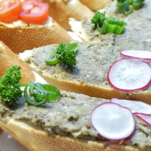 Ein schräg geschnittenes Sandwichbrötchen mit rustikalem Linsenaufstrich, dekoriert mit drei Radieschenscheiben, Frühlingszwiebelringen und einer krausen Petersilie. Serviert auf einem Silbertablett belegt mit Tortenpapier.