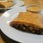 Ein Stück Sauerkraut-Erdäpfel Strudel angerichtet auf einem weißen Teller mit einer kleinen weißen Schale mit Paprika-Joghurt-Dip-