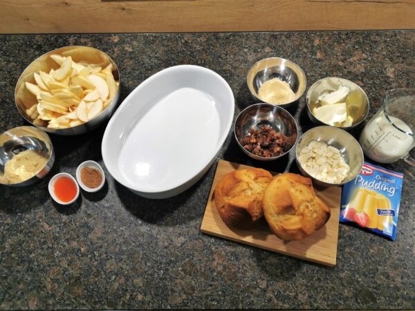 Alle vorbereiteten Zutaten für den Scheiterhaufen. Eine weiße Keramikauflaufform, eine Edelstahlschüssel mit geschälten Apfelscheiben, kleine Edelstahlschüsseln mit Rohrzucker, Vanillezucker, Margarine, Rosinen und Mandelblättchen, kleine weiße Schälchen mit Rum und Zimt, ein kleines Holzbrett mit zwei Osterpinzen darauf, ein Messbecher mit Pflanzenmilch und eine Packung Vanillepuddingpulver.