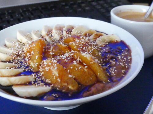 Schoko-Bananen Porridge mit Blutorangen angerichtet in einem weißen Teller, getoppt mit Dattelsirup und Poppies. Dazu serviert eine Tasse Kaffee.