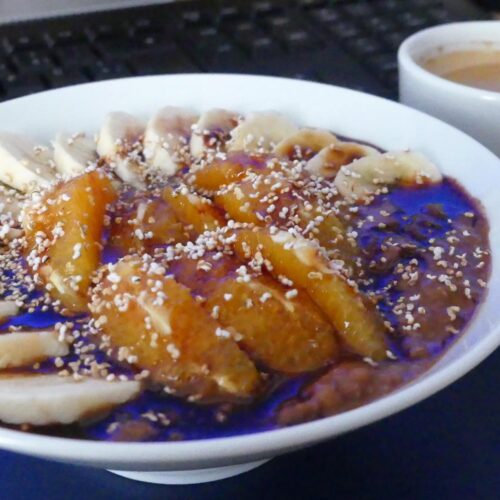 Schoko-Bananen Porridge mit Blutorangen angerichtet in einem weißen Teller, getoppt mit Dattelsirup und Poppies. Dazu serviert eine Tasse Kaffee.
