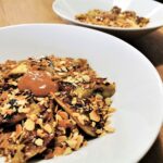 Schoko-Porridge mit karamellisierten Bananen und Mandeln serviert in einem weißen Teller, getoppt mit Mandelmus und Dattelsirup.