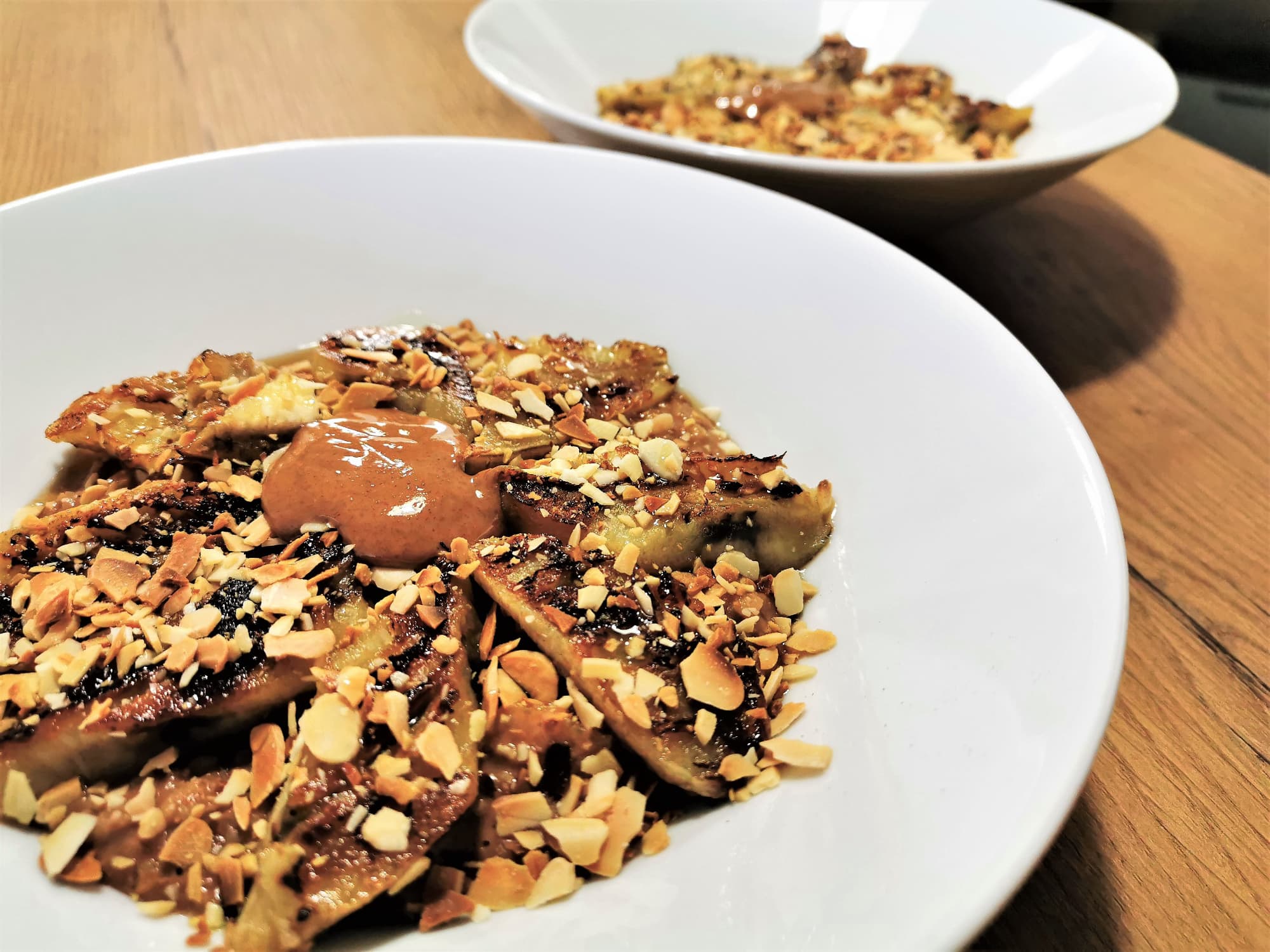 Schoko-Porridge mit karamellisierten Bananen und Mandeln serviert in einem weißen Teller, getoppt mit Mandelmus und Dattelsirup.