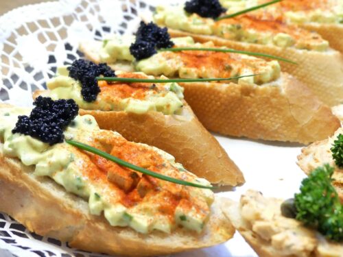 Ein schräg geschnittenes Sandwichbrötchen mit veganem Ei-Aufstrich, dekoriert mit zwei Schnittlauch, einem Klecks veganen Kaviar und fein bestreut mit Paprika. Serviert auf einem Silbertablett belegt mit Tortenpapier.