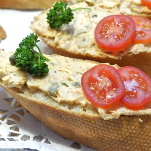 Ein schräg geschnittenes Sandwichbrötchen mit veganem Liptauer, dekoriert mit zwei Cocktailtomatenscheiben und einer krausen Petersilie. Serviert auf einem Silbertablett belegt mit Tortenpapier.