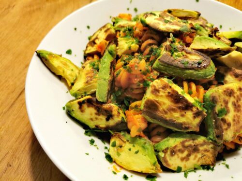 Fusilli mit Tomatensauce Arrabiata serviert in einem weißen Teller, dazu gebratene Avocadoscheiben in Stücke geschnitten und bestreut mit gehackter Petersilie.