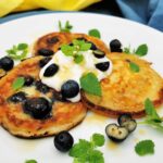 Drei Stück Heidelbeer-Pancakes angerichtet auf einem weißen Teller, getoppt mit einem Klecks Pflanzenjoghurt, Ahornsirup, frischen halbierten Heidelbeeren und Zitronenmelisse-Blättchen.