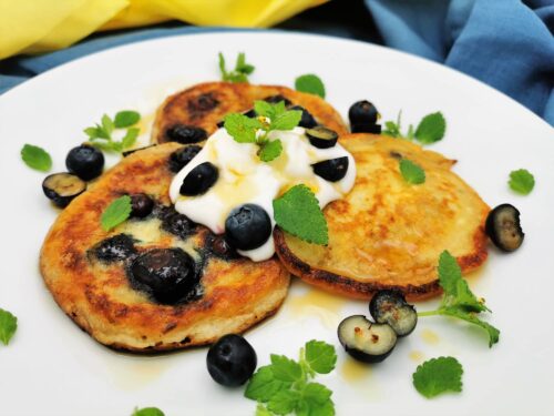 Drei Stück Heidelbeer-Pancakes angerichtet auf einem weißen Teller, getoppt mit einem Klecks Pflanzenjoghurt, Ahornsirup, frischen halbierten Heidelbeeren und Zitronenmelisse-Blättchen.