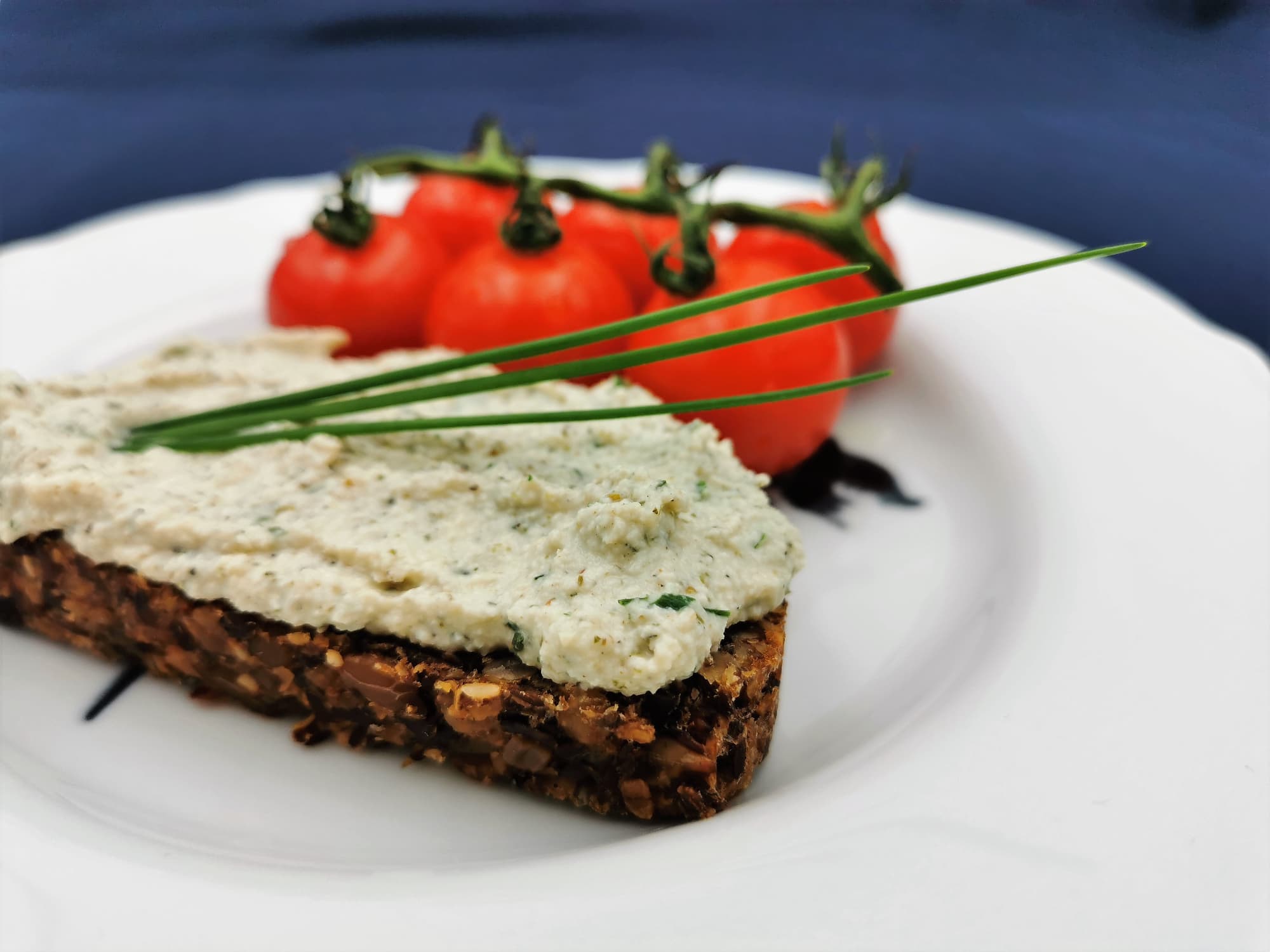 Eine Scheibe Körndlbrot mit Cashew-Kräuter-Frischkäse bestrichen, garniert mit Schnittlauchhalmen und einer kleine Rispe Cocktailtomaten, serviert auf einem weißen Teller.