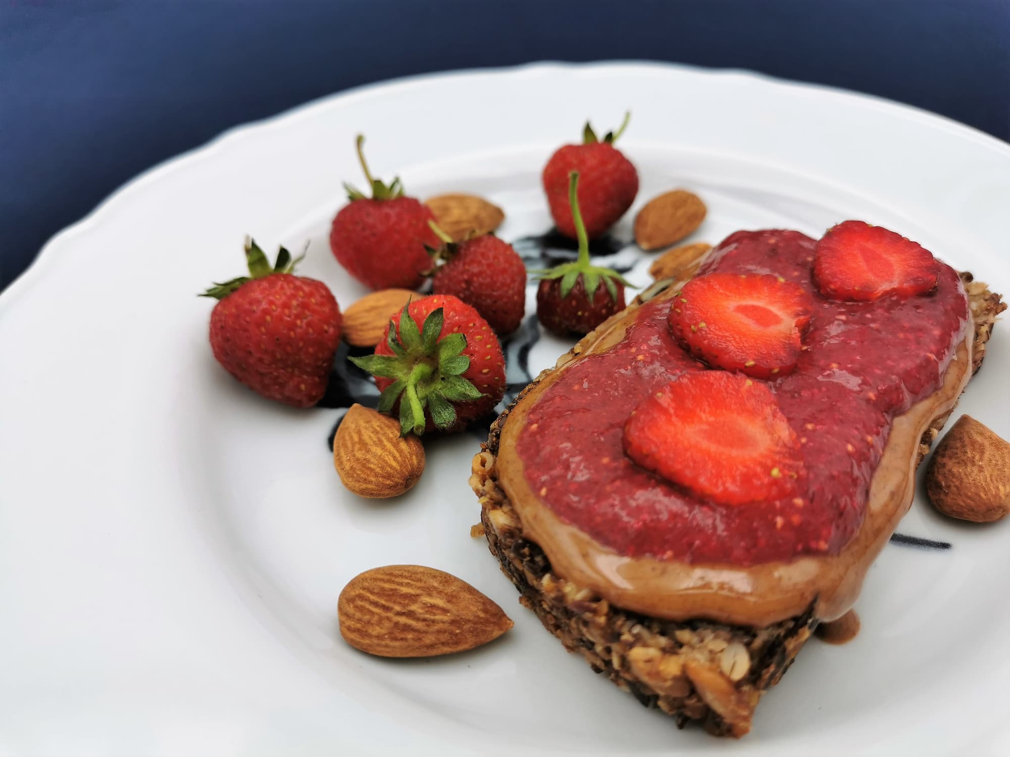 Eine Scheibe Körndlbrot mit Nussmus und Erdbeer-Chia-Jam, angerichtet auf einem weißen Teller, dekoriert mit frischen Erdbeeren und Mandeln.