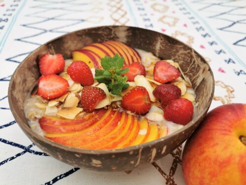 Eine Kokosnussbowl gefüllt mit Bananenjoghurt, belegt mit Nektarinenscheiben, Erdbeerhälften und gerösteten Mandelblättchen, garniert mit einem Zweig Zitronenmelisse.