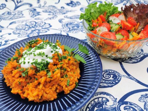 Reisfleisch serviert auf einem blauen Teller mit einem Klecks Pflanzenjoghurt und mit Petersilie bestreut. Daneben ein bunter Salat in einer gläsernen Salatschüssel.