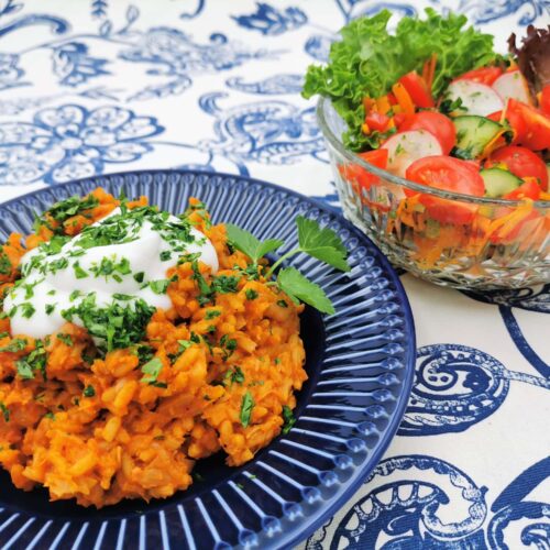 Reisfleisch serviert auf einem blauen Teller mit einem Klecks Pflanzenjoghurt und mit Petersilie bestreut. Daneben ein bunter Salat in einer gläsernen Salatschüssel.