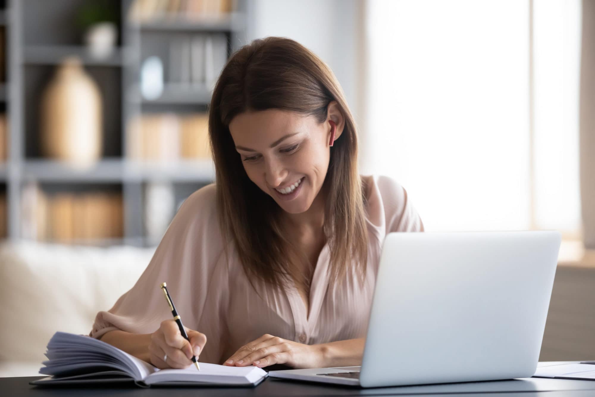 Smiling woman writing in notebook, using laptop at home