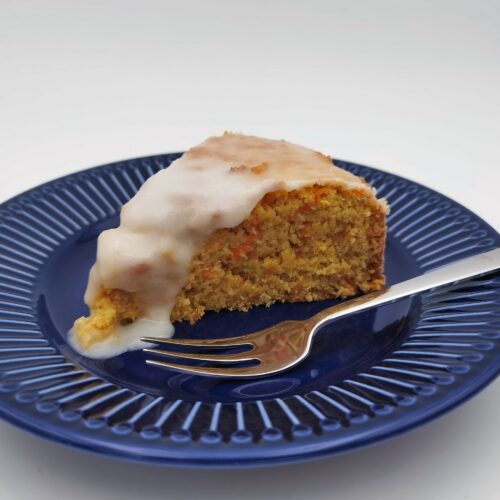 Ein Stück Karotten-Kokos-Orangen Kuchen mit verlaufendem Zuckerguss serviert auf einem blauen Desserteller mit einer Dessergabel.
