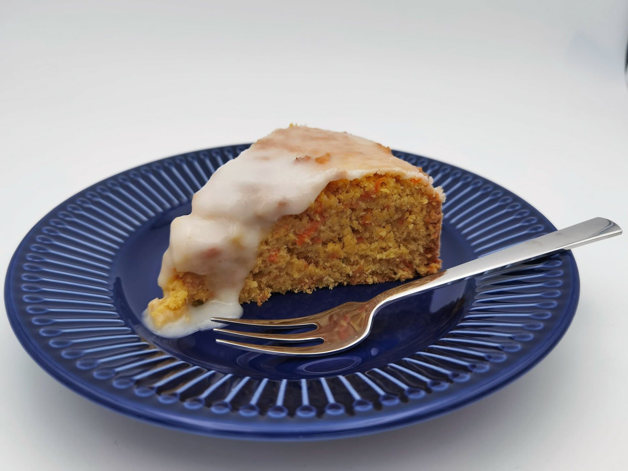 Ein Stück Karotten-Kokos-Orangen Kuchen mit verlaufendem Zuckerguss serviert auf einem blauen Desserteller mit einer Dessergabel.