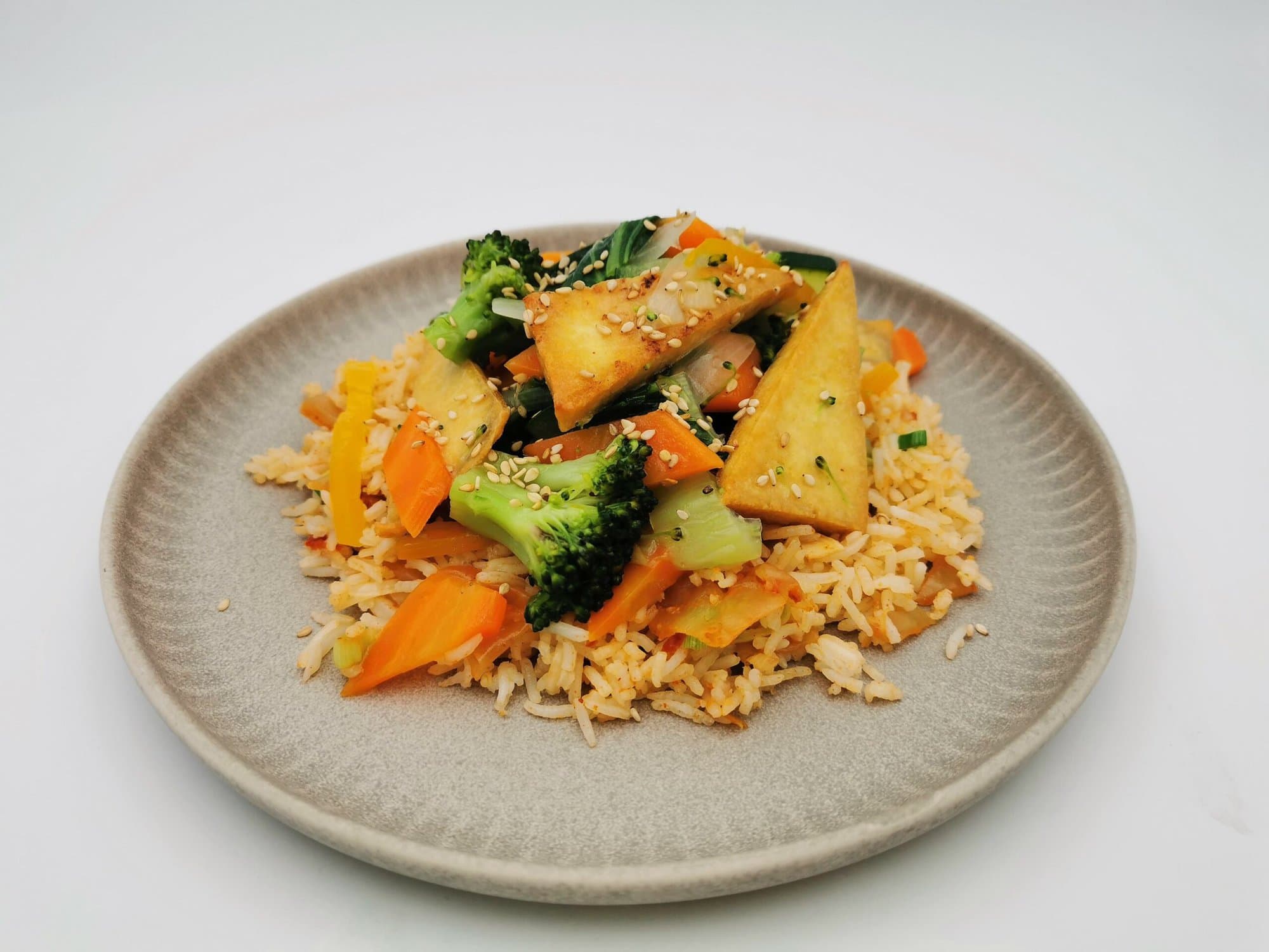Kimchi Fried Rice angerichtet mit gebratenem Wokgemüse bestehend aus Karotten und Brokkoli, mit gebratenen Tofuecken auf einem beigen Teller.