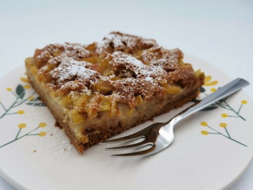 Ein Stück Rhabarberkuchen, bestreut mit Staubzucker, serviert auf einem Teller mit Blumendeko und einer Dessertgabel.